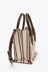 Bolso estilo bandolera con estampado a rayas verticales color marrón y crudo. Tiene asa de mano y asa fina extraíble con detalles metálicos. El bolso interior es a contraste de materiales con cierre mediante cremallera metálica. Cierre imantado.