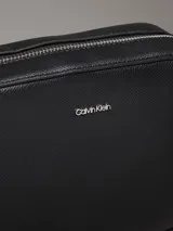 Bandolera negra Calvin Klein Must de piel sintética granulada, con correa ajustable jacquard con logo, cierre de cremallera, bolsillo interior y logo de metal plateado brillante en la parte delantera.