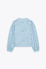Camisa celeste de cuello redondo y manga larga, con bordados perforados y volantes. Cierre frontal con botones ocultos por solapa.
