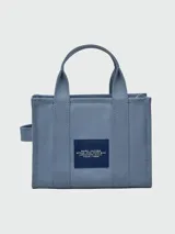 Bolso tote pequeño de lona color beige con estampado frontal en blanco con la leyenda "The Tote Bag Marc Jacobs". Tiene doble asa de mano y correa bandolera negra ajustable y extraíble.