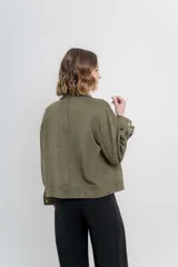 Chaqueta color beige de corte recto, estilo camisa, confeccionada en gamuza con elastano. Presenta cuello camisero, cierre frontal con botones metálicos, bolsillos grandes en el frente y charreteras en los hombros.