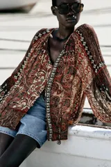 Blusa marrón con estampado de cachemir y detalles de hilo metalizado. Tiene cuello redondo con cordón ajustable y abalorios, escote en pico con nido de abeja, mangas largas con elástico en los puños y cierre frontal con botones forrados.