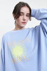 Buzo celeste de felpa con estampa de sol y texto "Golden Hour". Tiene escote redondo y hombros caídos.