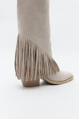 Bota texana de caña alta color beige confeccionada en gamuza, con punta fina, taco cuadrado de madera y detalle de flecos laterales. 