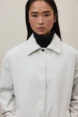 Chaqueta larga de color blanco roto, con cuello camisero y cierre frontal de broches. Presenta un corte holgado y bolsillos laterales.