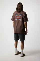 Remera marrón de corte recto y manga corta, con un parche bordado en el pecho con motivos geométricos en azul, rojo y blanco.
