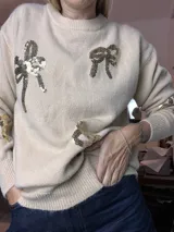 Suéter de punto color beige claro, con cuello redondo y puños y ruedo acanalados. Presenta apliques de moños hechos con lentejuelas doradas y plateadas en el frente y mangas.