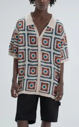 Camisa de manga corta tejida a crochet en punto abierto, con cuello tipo polo y cierre frontal con botones. Diseño con cuadrados de colores beige, naranja y azul. Corte oversize.