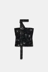 Top negro con escote recto y hombros descubiertos, con tiras tipo foulard para atar al cuello. Presenta bordados o aplicaciones de lentejuelas y abalorios en forma de flores blancas.