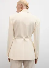 Blazer color crema con cuello de solapa, manga larga con puños abotonados, cinturón con hebilla y bolsillos delanteros con solapa.