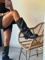 Bota de caña media-alta de cuero negro, estilo biker o motoquera, con caña arrugada y hebilla ajustable en el tobillo. Posee un taco bajo y suela gruesa.