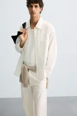 Camisa de corte relajado, color blanco hueso, con finas rayas verticales en tono grisáceo. Presenta cuello solapa, manga larga, bolsillo de parche en el pecho y bajo con aberturas laterales.