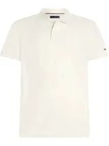 Chomba color crema con patrón integral de logos de la marca. Tiene cuello tipo polo, cartera de dos botones, mangas cortas y logo de Tommy Hilfiger bordado en la manga. Confeccionada en algodón BCI y elastolefín.