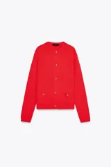 Chaqueta roja de punto con cuello redondo y manga larga. Tiene bolsillos delanteros de plastrón, cierre frontal con botones dorados con relieve y acabados en rib.