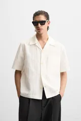 Camisa color blanco con textura de rayas verticales, corte relaxed fit, cuello bowling, manga corta, bolsillo de parche en el pecho y cierre frontal con botones.