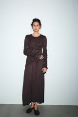 Vestido midi color marrón oscuro, con cuello redondo y manga larga. Presenta frunces diagonales en la parte superior delantera y en las mangas, y un bajo asimétrico.