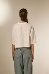 Remera blanca de manga 3/4, con cuello a la base y diseño holgado. Presenta un patrón de pequeños orificios y detalles de studs.