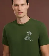 Remera verde militar de algodón texturizado, corte regular, cuello redondo y mangas cortas. Estampa tropical de una casa y palmeras en el pecho.
