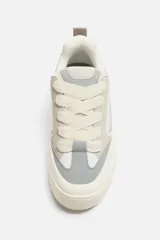 Zapatilla deportiva tipo skate con parte superior fabricada en combinación de piezas y materiales. Detalle de cordones maxi y suela con volumen. Color blanco, gris y crema.