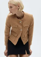 Chaqueta corta de tweed color marrón con cuello redondo, cierre frontal con botones metálicos, cuatro bolsillos de parche y terminaciones deshilachadas.