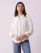 Blusa blanca de gasa con cuello a la base, escote en V y abertura central con botones. Tiene mangas 3/4 amplias con apliques de crochet y detalles plisados en la parte delantera.