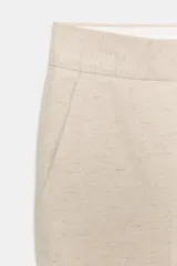 Pantalón tobillero color beige, de tiro medio, confeccionado en hilatura con algodón y lino. Presenta bolsillos laterales, raya planchada y cierre frontal con cremallera, botón y ganchos metálicos.