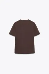Camiseta marrón de algodón de manga corta y cuello redondo. Presenta un bolsillo en el pecho con un broche metálico dorado con diseño intrincado.