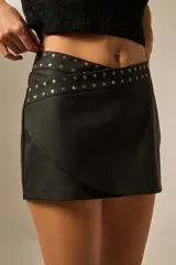 Minifalda negra de poliuretano con diseño cruzado y detalle de tachas plateadas en la cintura.