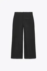 Pantalón negro de tiro alto con trabillas, bolsillos laterales y pinzas. Cierre frontal con cremallera, botón interior y gancho metálico.