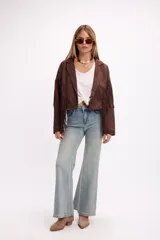 Chaqueta de efecto gamuza en color tostado, con solapas amplias, cierre frontal con un botón y flecos largos decorativos en la parte frontal y trasera de las mangas. Presenta una estructura definida y forro interior.