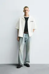 Camisa relaxed fit color blanco crudo, confeccionada en tejido de algodón con estructura. Cuello bowling y manga corta. Estampado frontal con la frase "ONLY DUST IS FOREVER" en negro. Cierre frontal de botonadura.