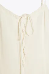 Vestido corto color crudo, de gasa, con escote en V y tirantes finos regulables. Presenta cordón cruzado con lazada en el delantero y falda con volantes. Cierre trasero con cremallera oculta.
