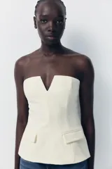 Top blanco de escote recto con detalle de pieza metálica en forma de V forrada y hombros descubiertos. Tiene falsos bolsillos con solapa en el delantero, forro interior combinado y cierre lateral con cremallera oculta en la costura.