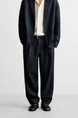 Pantalón de denim azul oscuro, corte wide fit, largo cropped, con pliegues frontales, bolsillos laterales y traseros asimétricos. Cierre frontal con cremallera y gancho metálico oculto.