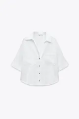 Camisa cropped de lino color blanco, con cuello solapa, escote pico, manga corta y dos bolsillos delanteros de parche. Cierre frontal con botones.