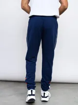 Pantalón de jogging azul marino con puños ajustados, cintura elástica con cordón y bolsillos laterales. Presenta una franja blanca con detalles en rojo a lo largo de la pierna, el escudo del Club Nacional de Football y el logo de Umbro.