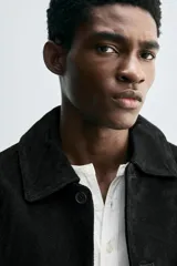 Cazadora de corte cropped fit, confeccionada en piel con acabado serraje (gamuza) de color negro. Presenta cuello solapa, manga larga, dos bolsillos de plastrón en la cadera y cierre frontal de botonadura.