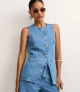 Chaleco femenino de jeans, sin mangas, con cuello redondo, diseño cinturado y botones dorados frontales. Presenta un corte asimétrico en el ruedo con solapas superpuestas y bolsillos de parche en la parte inferior.