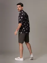 Camisa de manga corta con estampado de flores y hojas en blanco y gris sobre fondo negro. Tiene cuello clásico, cierre con botones y bolsillo de parche en el pecho.