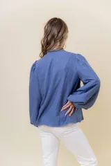 Camisa azul de calce relajado con bordado floral blanco en el frente. Presenta mangas largas amplias con ligero fruncido en el hombro y cierre frontal con botones.