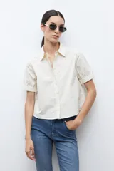 Camisa de algodón color beige claro, con cuello camisero, botones frontales y mangas cortas abullonadas con elástico en el puño.