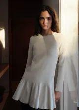 Vestido corto color crudo de tejido de punto fino con diseño de tablas en la falda, cuello redondo y manga larga.