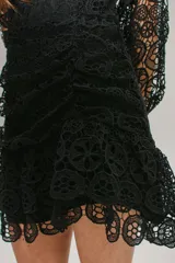 Vestido corto de encaje negro con escote en V, mangas largas abullonadas y falda con volados. Presenta cierre invisible en la espalda.