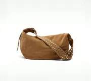 Bolso marrón de gamuza con forma de media luna y tachas plateadas. Tiene correa de hombro ajustable con hebilla metálica y cierre superior con cremallera.