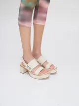 Sandalias de cuero color crema con plataforma y taco ancho. Presentan dos tiras anchas sobre el empeine con argollas metálicas decorativas y una tira trasera con elástico.