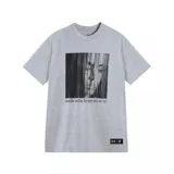 Remera de algodón gris melange, corte oversize, con cuello redondo. Estampado frontal en blanco y negro con el rostro de una mujer parcialmente cubierto por líneas verticales y el texto "nadie sabe lo que no se ve" en la parte inferior. Incluye un tag negro con logos de Under Armour y Tini en la parte inferior.