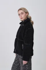 Campera de corderito color negro, con cuello alto y cierre frontal completo. Presenta detalles de vivos en cuero sintético en el cierre, bolsillos y costuras, además de una hebilla decorativa en el cuello.