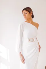 Vestido largo blanco de seda, con un hombro descubierto, manga larga abullonada y falda irregular con drapeado lateral. Incluye cinturón blanco con hebilla de alpaca y nácar.