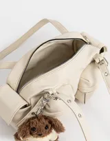 Cartera bandolera color crema con múltiples bolsillos frontales y laterales, doble asa corta regulable y charm de oso de peluche.