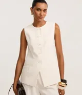 Chaleco largo color crema, con cuello redondo, sin mangas, cierre frontal con botones forrados, bolsillos delanteros falsos y corte recto.